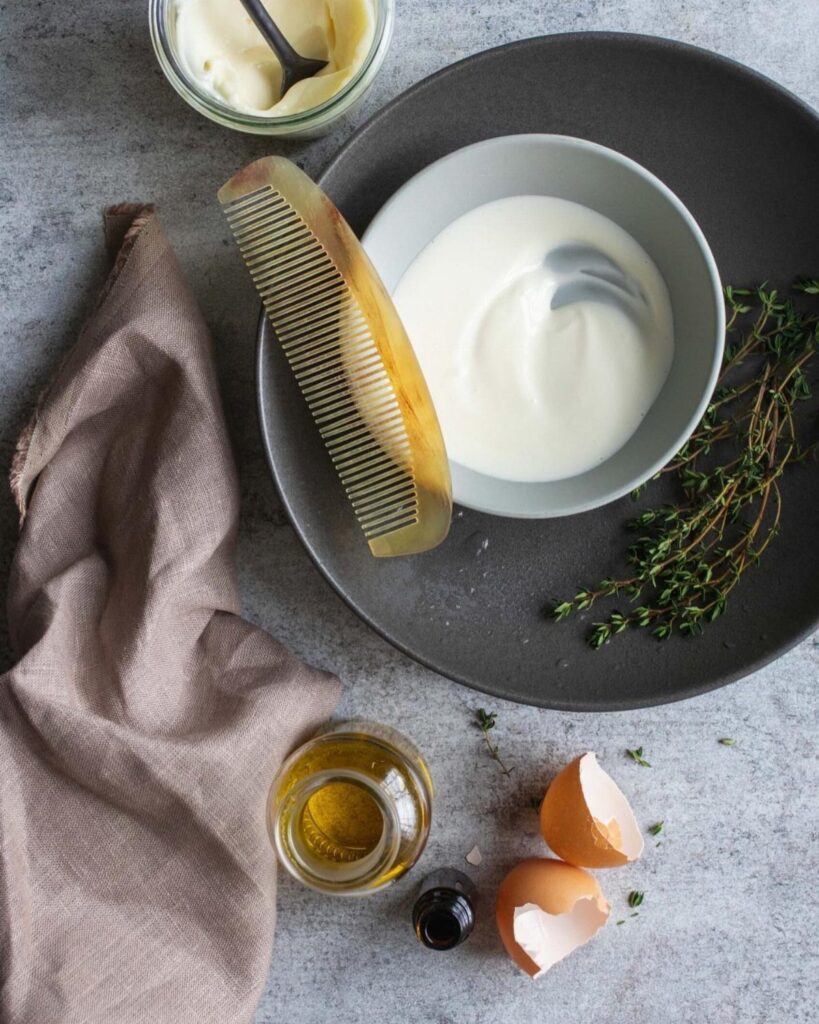 Bowl of yogurt, eggshells, oil, and mayonnaise with a comb on grey surface.