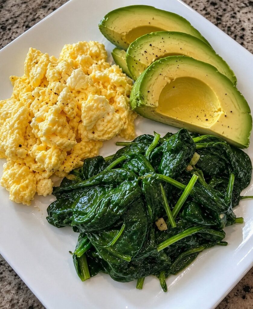 Scrambled eggs, sliced avocado, and sautéed spinach served on a white square plate.