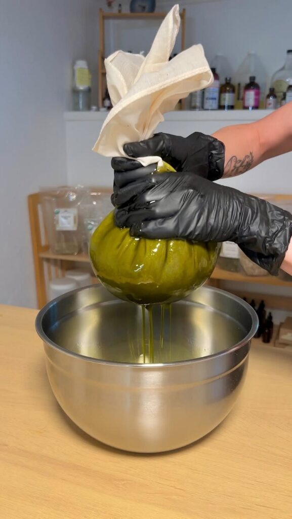 Hands in black gloves squeezing herbal oil from a cloth bag into a bowl.