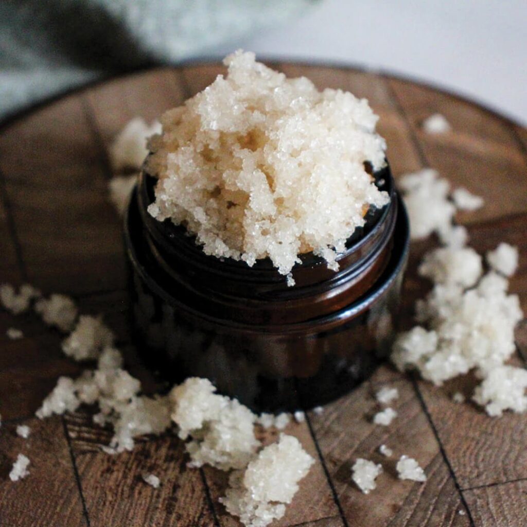 White crystalline scrub overflowing from a dark brown glass jar on wooden surface
