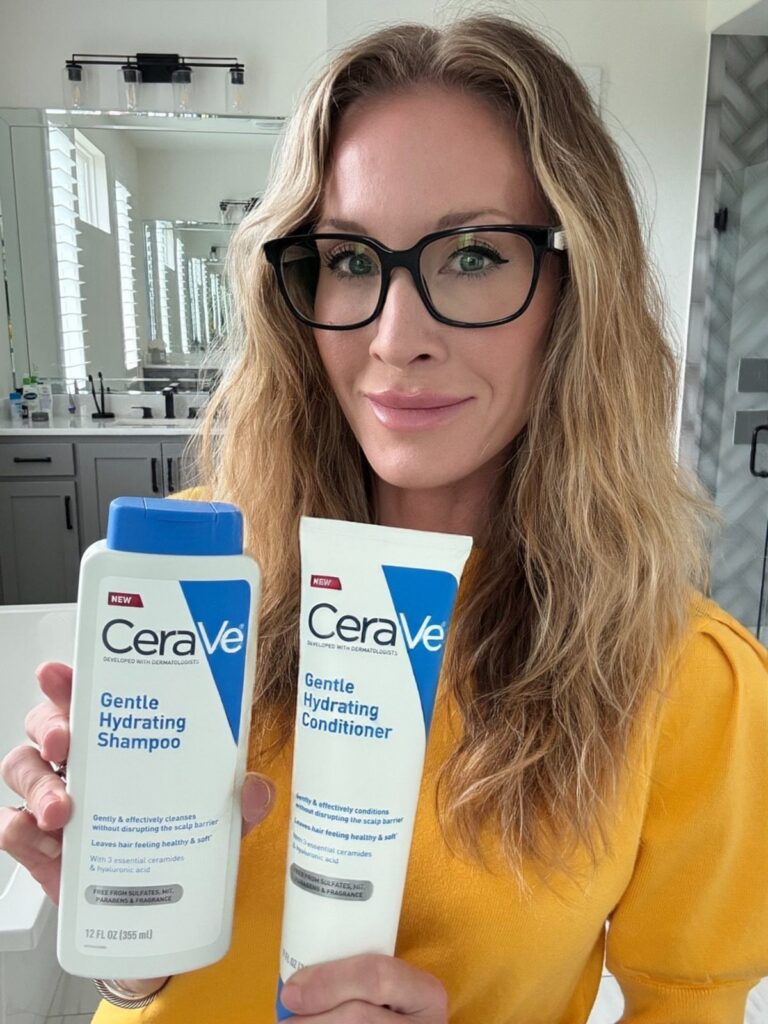 Woman holding CeraVe Gentle Hydrating Shampoo and Conditioner bottles in a bright bathroom.